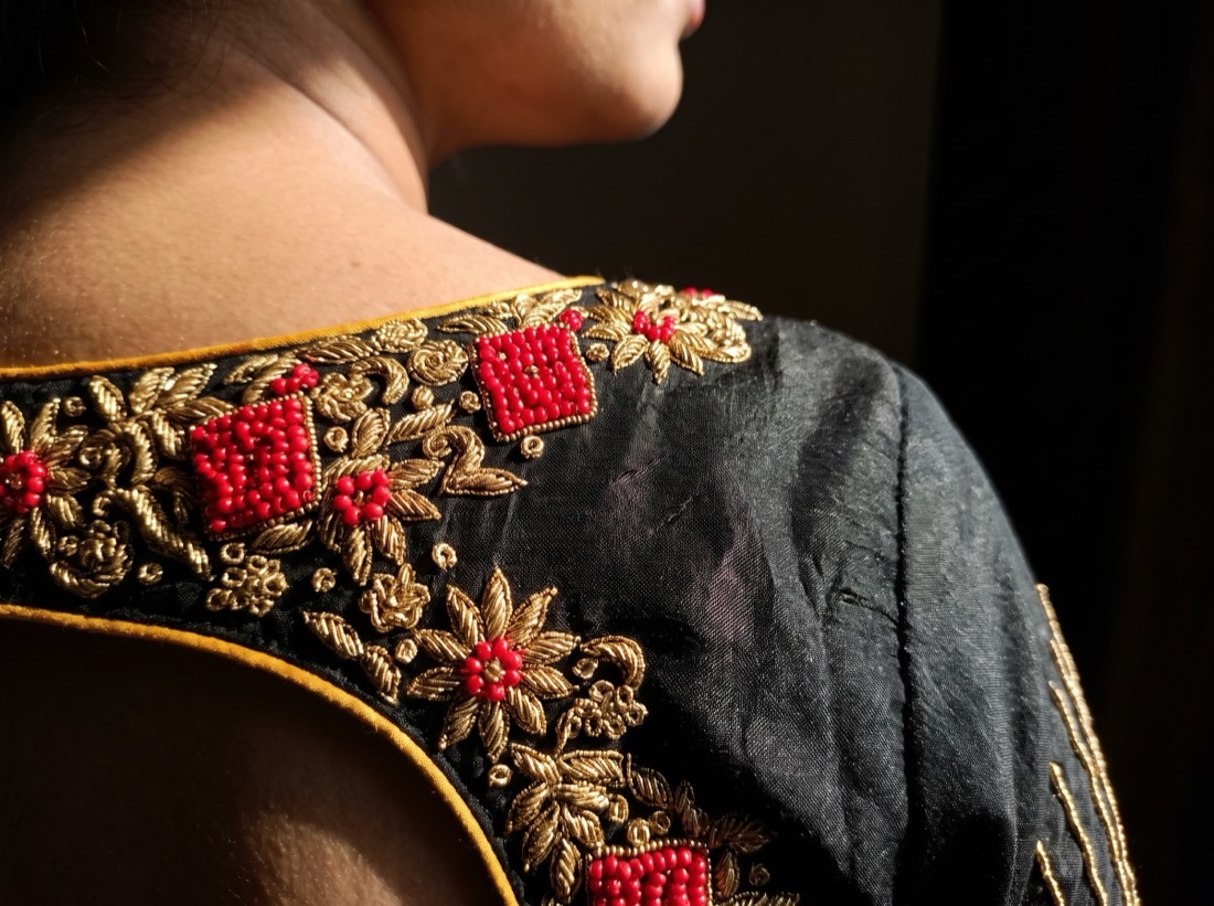 Macro close-up of the shoulder of a black blouse: raised gold zardosi coil-work flowers, clustered red bead squares and fine mustard piping.
