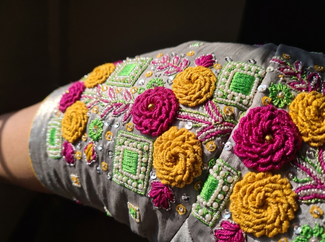 Macro detail of raised magenta and mustard aari rosettes with pearl-outlined green squares on grey silk.