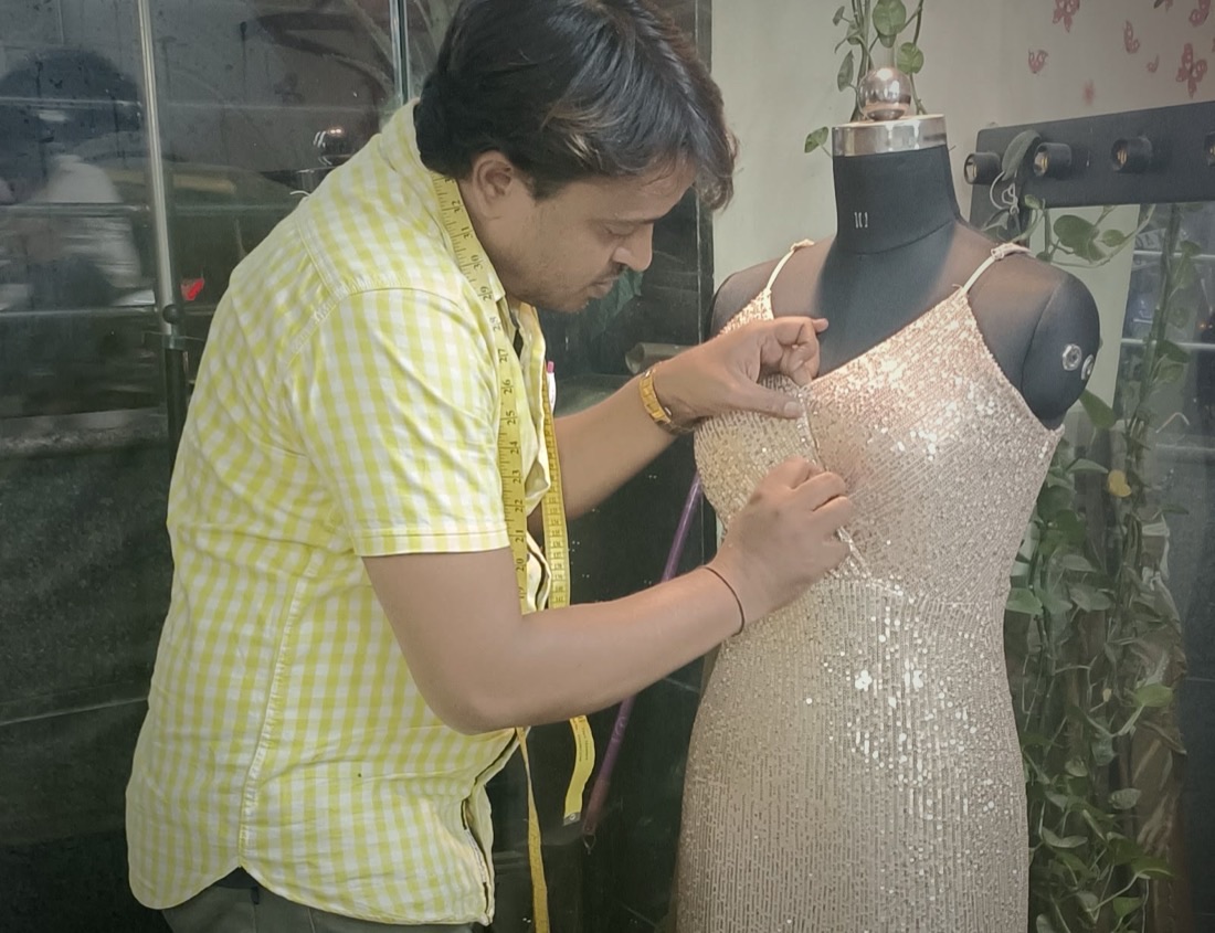 Inside the studio: the tailor fitting a sequinned gown onto a dress form, with rattan pendant lamps and rolls of fabric behind him.