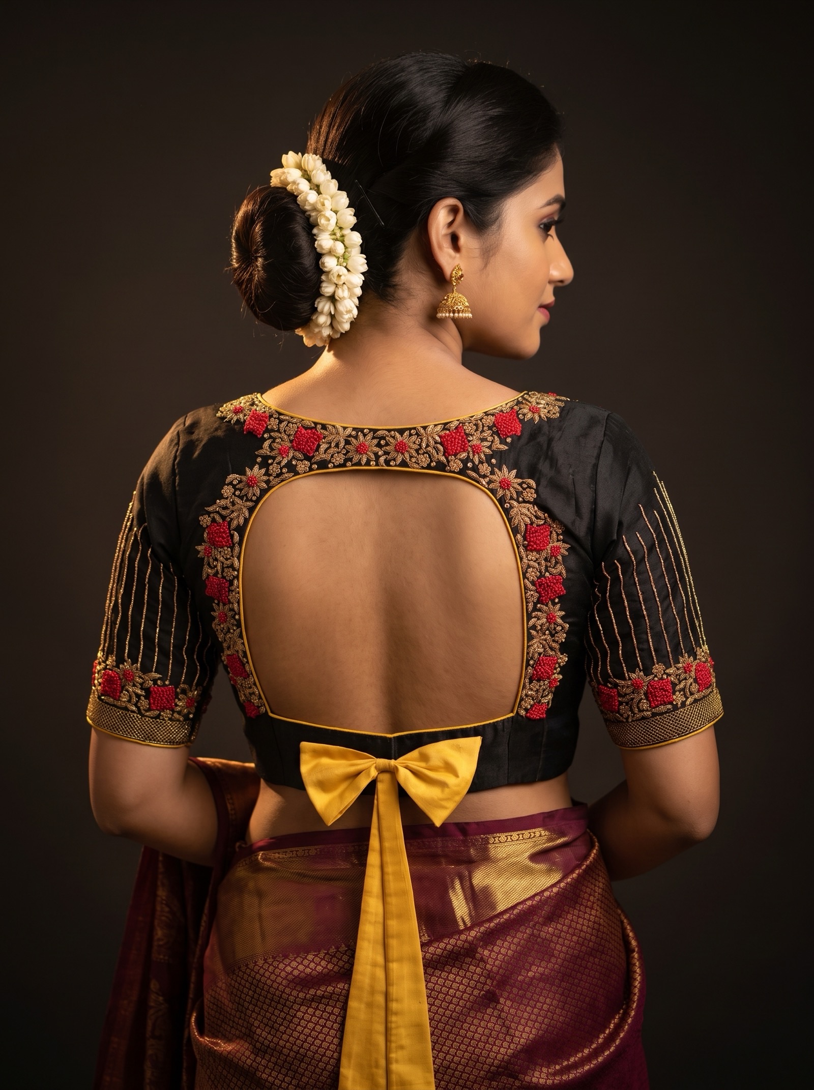 A woman seen from behind wearing a black silk blouse with gold zardosi scrollwork, red bead clusters and a mustard silk bow tied at the back.